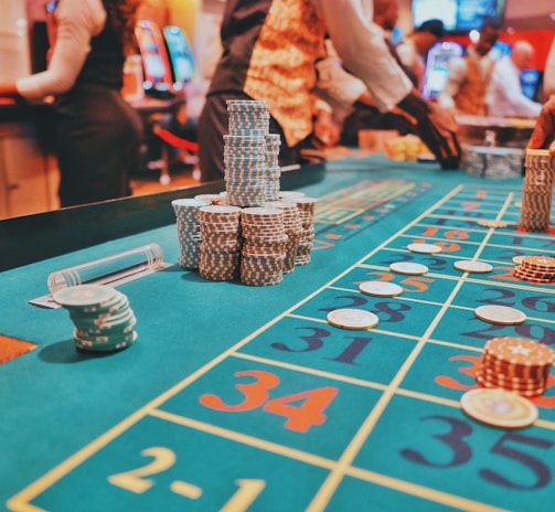 A casino scene with a roulette table features prominently, covered in green felt with colorful numbers and betting squares. Several stacks of poker chips are placed on different numbers, and the hands of several people are visible as they interact with the table. In the background, there are slot machines and more people, creating a vibrant and busy atmosphere.