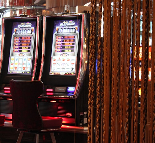 A pair of casino slot machines, each with detailed screens displaying various game outcomes and jackpot amounts. The chairs in front of the slot machines are dark and cushioned for comfort. The background features a hanging curtain made of golden ropes and other casino decorations are visible.