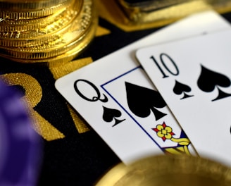 A close-up view of playing cards, specifically a Queen and a Ten of spades, surrounded by shiny gold coins and part of a poker chip, suggesting a gambling or casino theme.