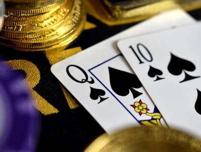 A close-up view of playing cards, specifically a Queen and a Ten of spades, surrounded by shiny gold coins and part of a poker chip, suggesting a gambling or casino theme.