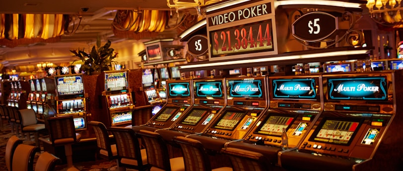 A casino floor featuring rows of brightly lit video poker machines. The area is illuminated by elegant chandeliers, with a display board showing a large sum of money, suggesting a possible jackpot. The rich decor includes luxurious carpets and comfortable seating, adding to the opulent atmosphere.