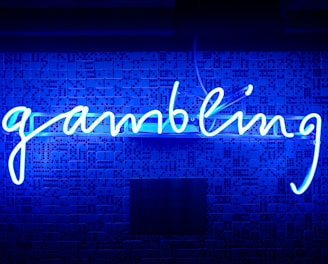 A bright blue neon sign spells out the word 'gambling' in cursive script. The background features a textured wall covered in domino tiles, also illuminated by the blue light, creating a vivid and visually striking scene.