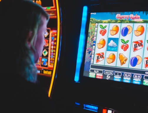 A person is closely observing a digital slot machine with colorful fruit symbols, bars, and a bell. The slot machine screen displays symbols like oranges, plums, cherries, and clouds. The background is dimly lit, creating a focused and somewhat intense atmosphere.