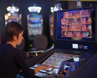 A person is seated at a slot machine console in a casino, focusing intently on the game displayed on the screen. The background shows several other illuminated gaming machines, adding a vibrant and colorful atmosphere to the scene.