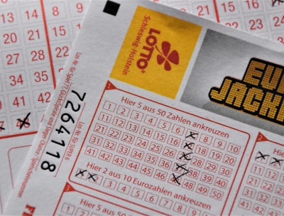 A close-up image of lottery tickets featuring crossed-out numbers, brand logos, and various numbers in a grid format. The tickets have sections for different games, and numbers are marked with black crosses.