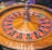 A roulette wheel with alternating red and black numbered pockets on a wooden platform. The wheel contains a small white ball resting in one of the pockets, indicating gameplay. Surrounding the wheel are stacks of colorful poker chips, suggesting a casino environment.