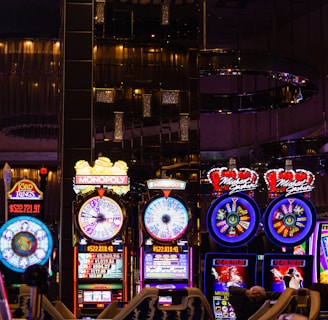 A lively casino scene featuring a row of bright, colorful slot machines with themes like Monopoly, Michael Jackson, and Lord of the Rings. The gaming machines display a variety of flashing lights and digital screens, creating an energetic and exciting atmosphere. Reflections of the lights are visible in glossy surfaces above the machines.
