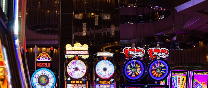 A lively casino scene featuring a row of bright, colorful slot machines with themes like Monopoly, Michael Jackson, and Lord of the Rings. The gaming machines display a variety of flashing lights and digital screens, creating an energetic and exciting atmosphere. Reflections of the lights are visible in glossy surfaces above the machines.