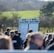A group of people gathered outdoors with two large betting boards in the background labeled 'REG DAVIES'. The setting appears to be a countryside location with trees and fields visible in the distance. The crowd consists of individuals dressed in jackets and hats, engaging in conversation.