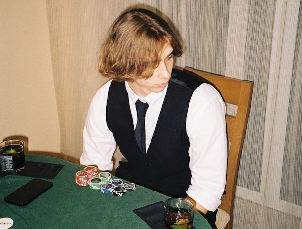 A person with shoulder-length hair is sitting at a poker table. They are wearing a white shirt, black vest, and tie, with an array of poker chips in front of them. There are two glasses with drinks on the table, and a smartphone is placed nearby. The background consists of vertical striped curtains.