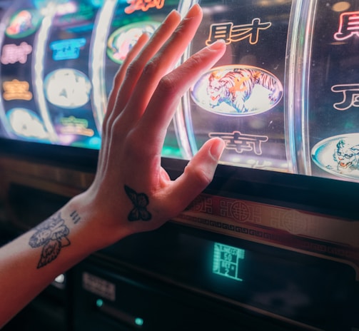 A hand with tattoos reaches towards a brightly lit slot machine, featuring various colorful symbols including a tiger. The image captures the moment of anticipation as the person engages with the machine during a game of chance.