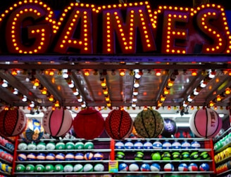 A brightly lit carnival game stall with a large 'GAMES' sign at the top surrounded by red and yellow lights. Various sports balls, including basketballs and soccer balls, are hanging from the ceiling and neatly arranged on shelves below.