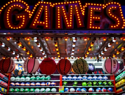A brightly lit carnival game stall with a large 'GAMES' sign at the top surrounded by red and yellow lights. Various sports balls, including basketballs and soccer balls, are hanging from the ceiling and neatly arranged on shelves below.