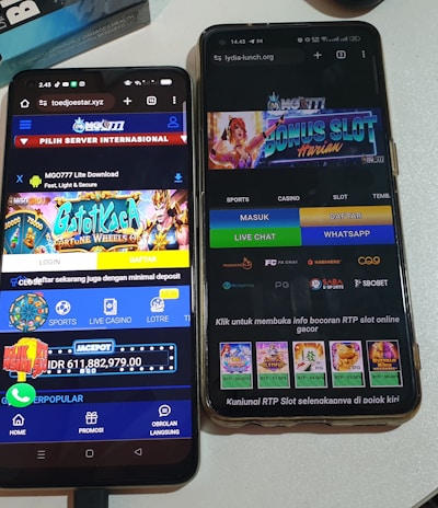 Two smartphones are displayed on a white surface, both showing gambling websites with colorful graphics, text, and various options for logging in and accessing games. The screens are bright and filled with promotional content related to casino and slot games.